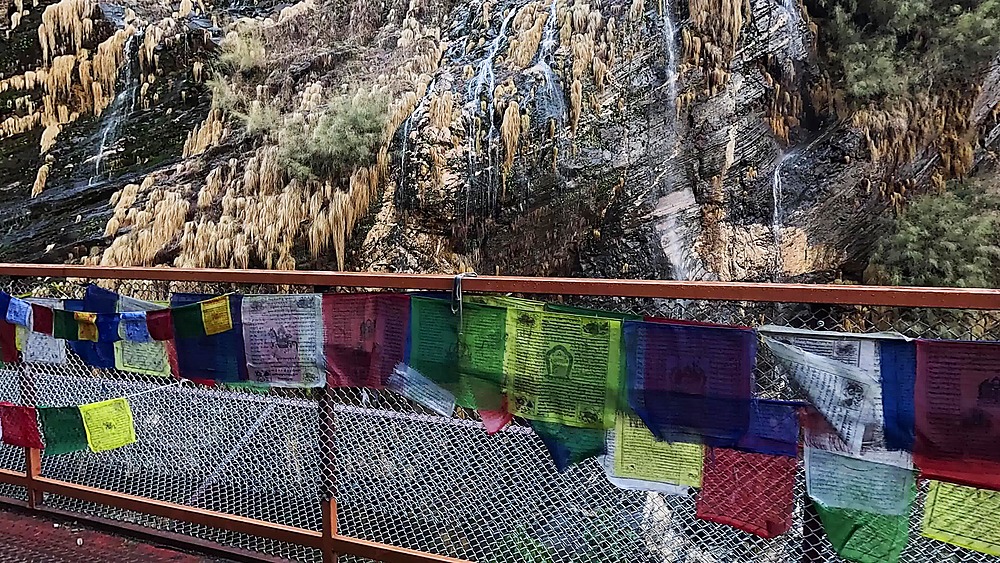 Colourful Buddhist prayer flags on a bridge railing below a waterfall cliff, Annapurna, Nepal