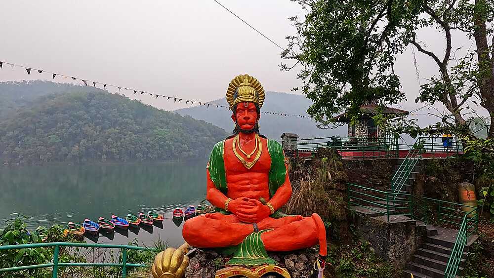 Hanuman statue in red and green at Shiva Mandir Gaurighat, overlooking Phewa Lake with boats below