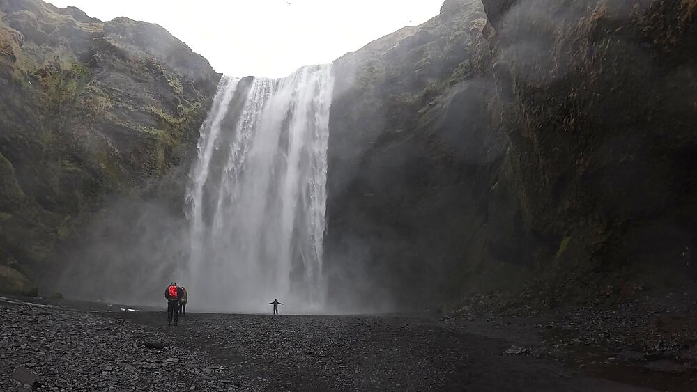 Skogafoss two silhouetted figures arms outstretched in spray mist directly before 60m waterfall, South Iceland
