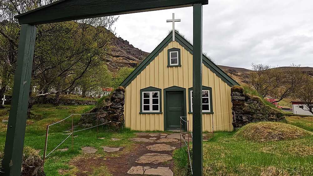 Hofskirkja historic turf church wide shot with gate, yellow facade green trim and turf roof, Oraefi, Iceland