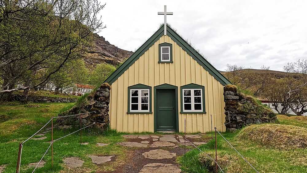 Hofskirkja turf church frontal view, yellow facade green door stone walls turf roof and cross, Oraefi, Iceland
