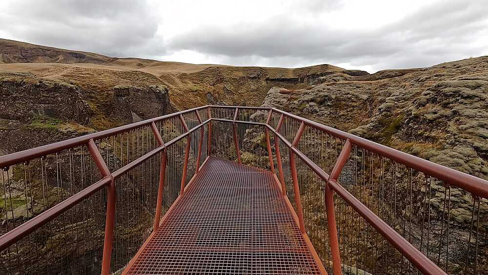 Red metal viewing bridge over Fjadrargljufur canyon gorge, moss-covered palagonite walls both sides, Iceland