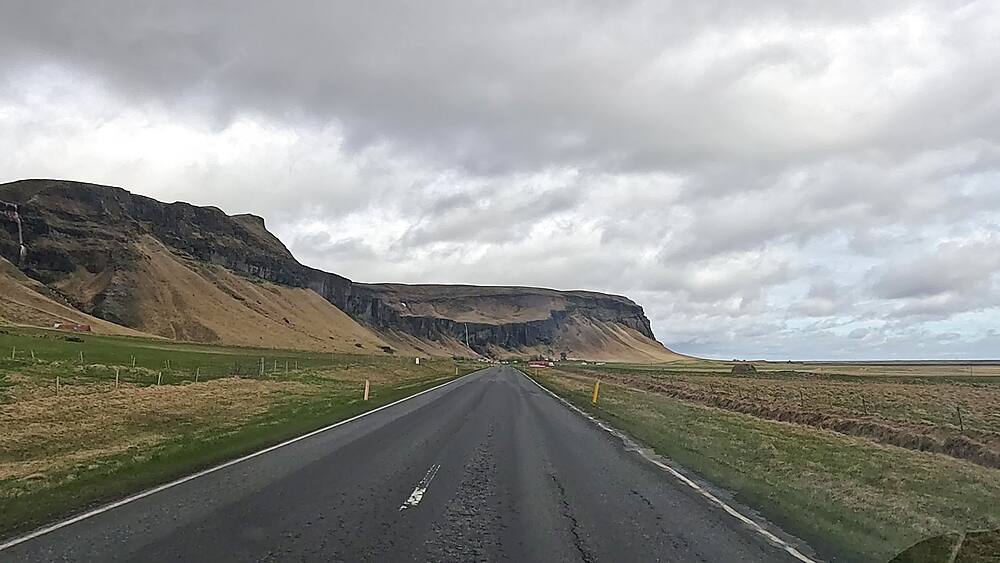 Dashcam POV on straight Route 1 South Coast road toward flat-topped cliff escarpment, South Iceland