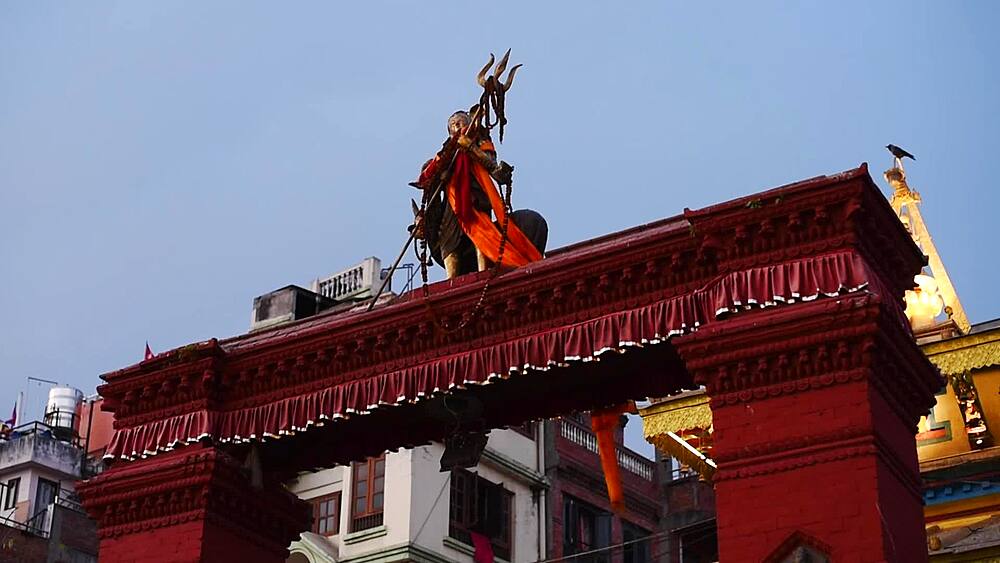 Trident-bearing Shiva statue crowns the red gateway of Mahendreshwor Temple at dusk, a crow perched alongside, Kathmandu, Nepal