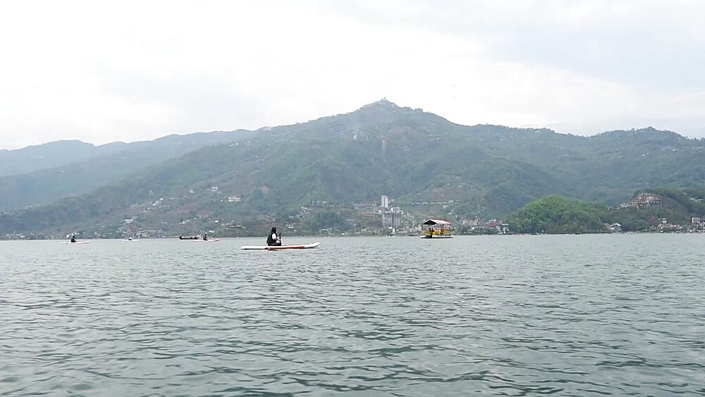 Stand-up paddleboarder glides across Phewa Lake, tracked from a boat, World Peace Pagoda on the forested ridge, Pokhara, Nepal