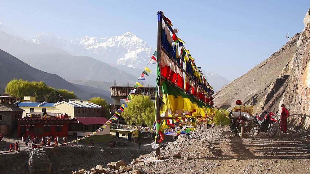 Prayer-flag pole above Kagbeni village with a snow peak beyond, riders on the dusty track