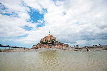 Mont Saint Michel seen in shallow tidal waters with visitors walking along the path in Normandy France