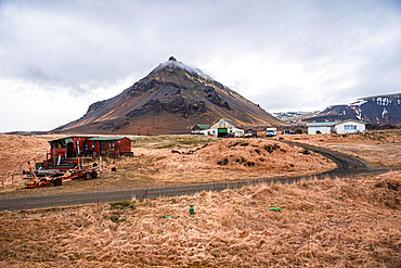 A sharply pointed volcanic mountain rises above a rural farmstead and flat autumn plains near Arnarstapi, Sn&aelig;fellsnes Peninsula, West Iceland, under a heavy overcast sky.