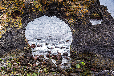 The perfectly circular Gatklettur sea arch frames a view of crashing Atlantic waves and wet barnacled rocks below, Arnarstapi, Sn&aelig;fellsnes Peninsula, West Iceland, the ancient basalt worn smooth by centuries of ocean swell.