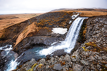 Sv&ouml;&eth;ufoss waterfall seen from above dropping into a rocky canyon with snow-patched moorland, near Rif, Sn&aelig;fellsnes Peninsula, West Iceland