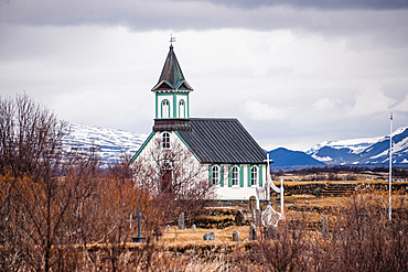 Historic white timber church with green steeple against snow-capped mountains in Thingvellir National Park, Iceland
