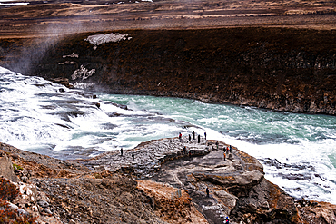 Gullfoss, Golden Circle, South Iceland. Tiny figures on a rocky outcrop dwarfed by the turquoise glacial torrent rushing through the canyon. Scale of nature, iconic waterfall and dramatic scenery on the Hv&iacute;t&aacute; river.