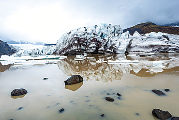 Sv&iacute;nafellsj&ouml;kull glacier terminus, Vatnaj&ouml;kull National Park, South Iceland. Stranded ice and the crevassed front reflect across a still lagoon strewn with dark boulders under a bright cloudy sky. Reflection, meltwater, glacial wilderness.