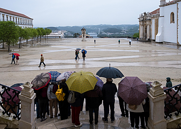 Visitors gather under umbrellas during rainfall in the courtyard of the University of Coimbra in Portugal, observing the surroundings