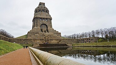 The massive monument is reflected in the water, flanked by a path and bare trees, Monument to the Battle of the Nations, Leipzig, Germany