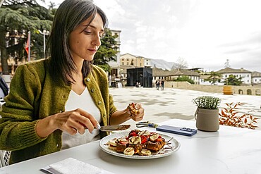 Close up woman sit in cafe enjoying large, indulgent waffle dessert portion topped with fruit and chocolate at an outdoor cafe. Travel lifestyle dining in historic stone town square of Gjirokastra