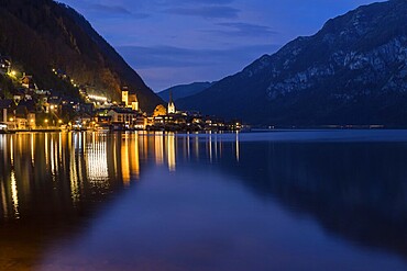 The enchanting village of Hallstatt, surrounded by majestic mountains, is beautifully illuminated at night The calm lake reflects the lights, creating a serene atmosphere