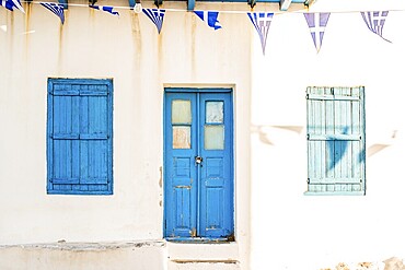 Sunlit Greek building facade featuring blue wooden doors and shutters in Plaka, Milos island. Greek flags hang vibrantly above, capturing coastal charm and Mediterranean aesthetic