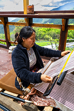 Woman weaves traditional clothes on a loom in Eastern Bhutan while surrounded by nature and mountains in the background