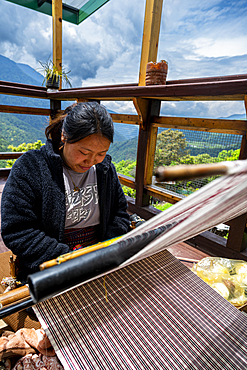 Woman weaves traditional clothes on a loom in Eastern Bhutan while surrounded by a mountain view and natural scenery