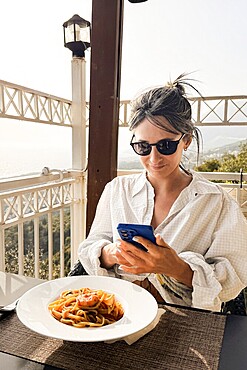 Woman taking photo eating shrimp spaghetti on scenic Mediterranean restaurant terrace. Golden hour lighting, coastline views. Delicious seafood healthy fresh traditional eating on summer holiday