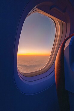 A serene sunset visible through the oval window of an airplane, showcasing a vast cloudscape tinged in hues of orange and blue