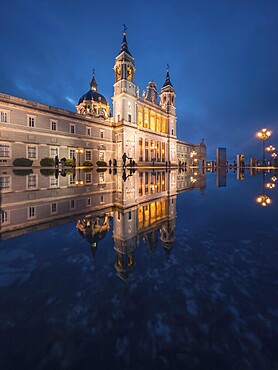 The majestic Almudena Cathedral of Madrid illuminated at dusk, its ornate architecture mirrored beautifully in the water. The serene ambiance and glowing lights create a breathtaking scene