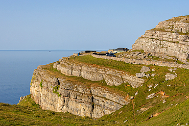 A scenic view of a rugged cliffside path overlooking the sea, with a house and vehicles in the distance at Great Orme, in Llandudno, North Wales.