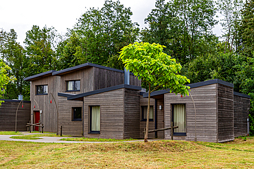 A contemporary wooden cabin nestled among trees, featuring a dark roof and large windows with curtains at Center Parcs Villages Nature Paris, France.