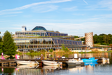 A contemporary glass structure with a green roof sits beside a calm waterway with docked boats and a whimsical flamingo float at Center Parcs Villages Nature Paris, France.
