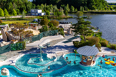 A scenic outdoor swimming pool with a lake and surrounding trees, featuring a water slide and a small gazebo at Center Parcs Villages Nature Paris, France.