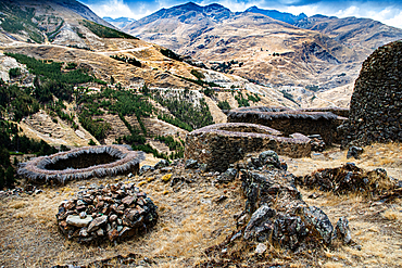 Ruins of grain towers overlook a remote valley in Peru surrounded by mountains and fields under a cloudy sky